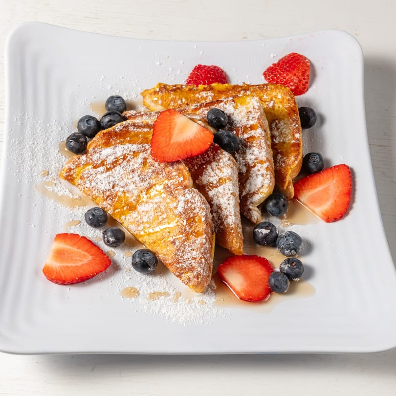 Golden French toast triangles, dusted with powdered sugar, drizzled with syrup, and garnished with fresh strawberries and blueberries.